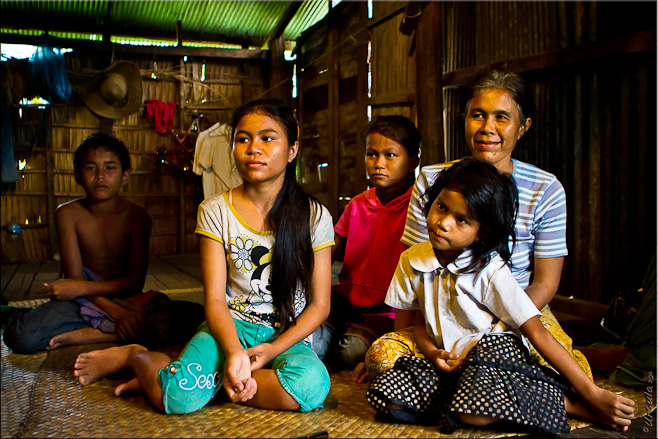 Khmer mother, three daughters and son seated on a grass mat in a bamboo house.