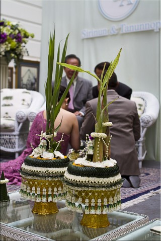 Foreground: Two elaborate bai sri (Thai floral decorations). Background: Newlyweds and their parents.