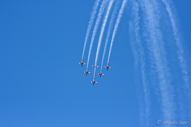 Six red and white PC-9/A ( two-seat single-engine turboprop aircraft) flying downward in triangle formation; vapour trails behind. 