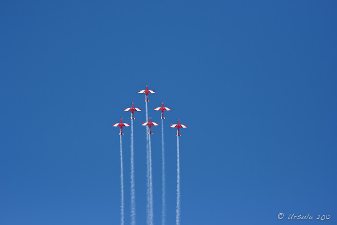 Six red and white PC-9/A ( two-seat single-engine turboprop aircraft) flying in triangle formation. 