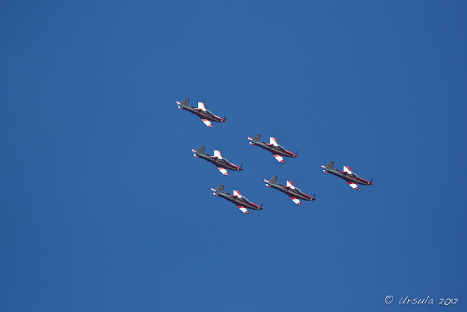 Six red and white PC-9/A ( two-seat single-engine turboprop aircraft) flying downward in triangle formation. 