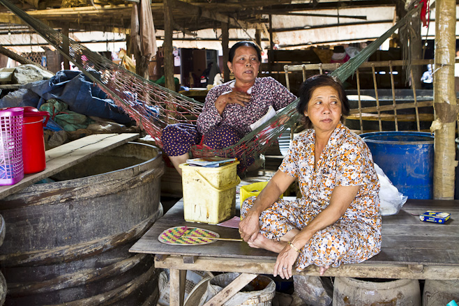 Two khmer women in cotton dresses sitting in a large warehouse-like building.