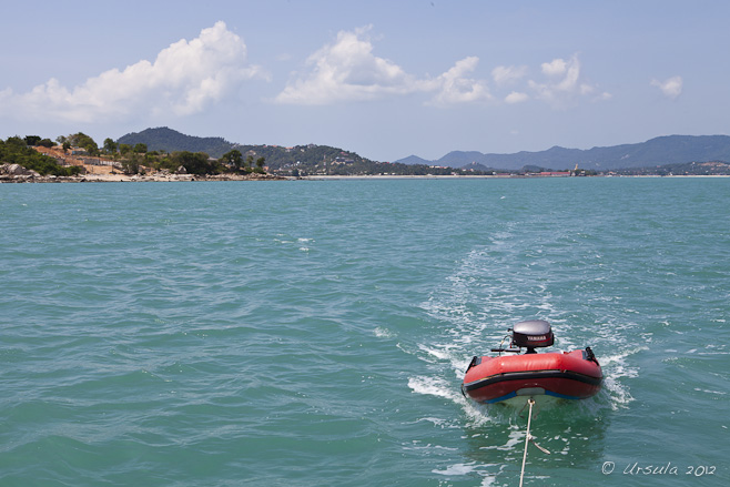 A red dinghy dragging behind a boat on turquoise waters. Mountains in the distance.