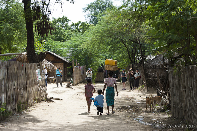 A woman with a load on her head walks into a Burmese village, while tourists photograph.