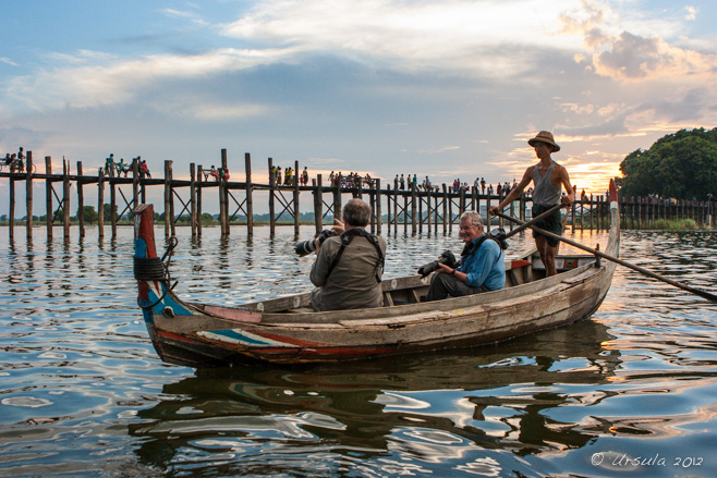 Standing boatman in a pith-hat and two men seated with large cameras, watching the foot-traffic on U Bein Bridge, Amarapura, Myanmar