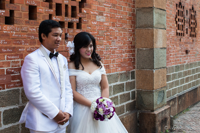 Vietnamese Wedding Couple in white in font of a red brick wall of Notre Dame Cathedral, Ho Chi Minh City Vietnam